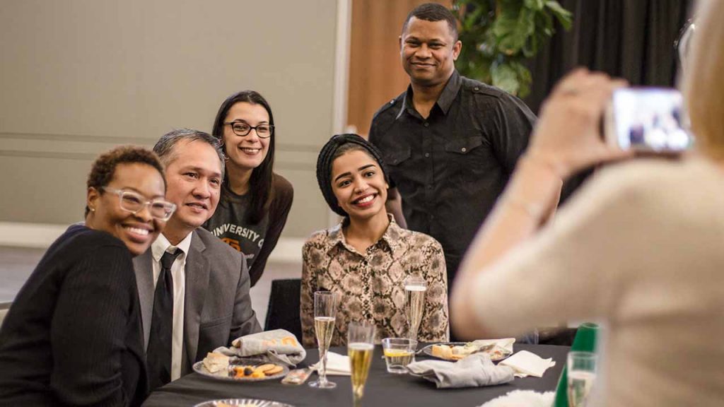 A group of alumni around a table pose for a photo at a Comets Take Flight event. Food and drinks are on the table in front of them.