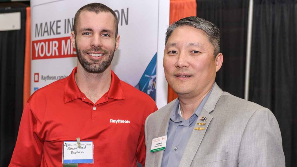 Two people at a career expo on campus. One is representing Raytheon and wearing a branded polo shirt. Both wearing name tags. 