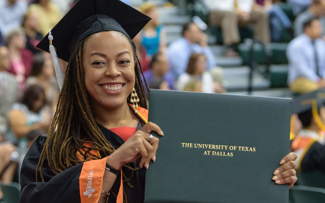 A graduate smiles while holding a diploma holder during commencement.