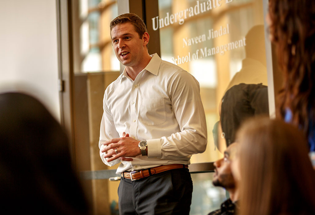 A person speaks to several others in front of a glass door that reads Undergraduate and Naveen Jindal School of Management. 